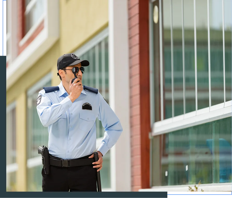 Male security guard in uniform, wearing sunglasses and a cap, holds a walkie-talkie near his mouth while standing in front of a building with large glass windows.